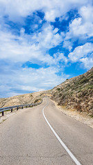 An empty and steep road going up the hill. Road is very curvy and dangerous. The slopes of the hill are barren, with some small bushes. Thick clouds on the sky. Road less travelled..