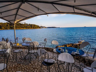 An elegant and nicely decorated restaurant located directly by the seashore. The tables are covered with blue tablecloth, chairs have white covers. In the back there is an island. Clear and sunny day