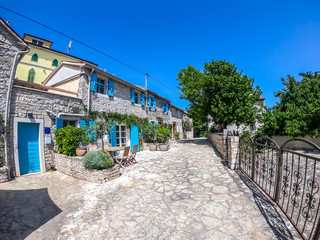 A view from a paved street on a little stony house with blue shutters. The view on the house is disturbed by the plants, growing next to it. There is a gate on the other side. Mediterranean culture.