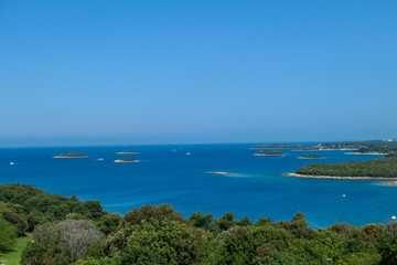 A view on the sea from a little hill. There are plenty small island around the coastal line, each of them overgrown with trees and bushes. There are few boats crossing the sea. Clear and sunny day.