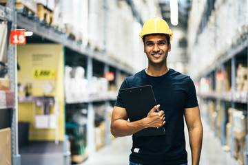 Smart Indian engineer man worker wearing safety helmet doing stocktaking of product management in cardboard box on shelves in warehouse. Factory physical inventory count.
