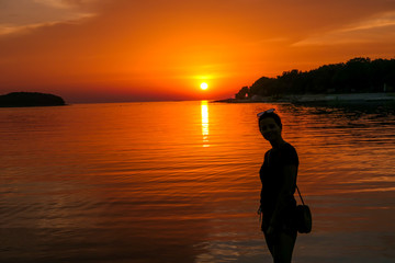 Fototapeta premium A girl enjoying the sunset by a beach. The sun sets over the horizon. The sun beams reflecting in the calm sea waters. There is an island on the side. The sky turns yellow. Girl enjoys the spectacle