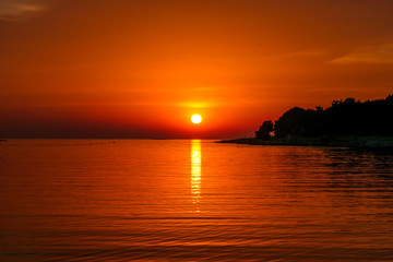 Fototapeta na wymiar Romantic sunset by a beach. The sun sets over the horizon. The sun beams reflecting in the calm sea waters. There is an island on the side. Few birds flying around. The sky turns yellow and orange