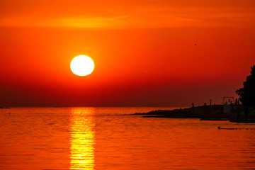 Romantic sunset by a beach. The sun sets over the horizon. The sun beams reflecting in the calm sea waters. There is an island on the side. Few birds flying around. The sky turns yellow and orange