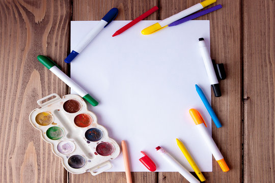 A white sheet of paper lay on a wooden table, near , pencils, paints and markers. Back to school. school and office supplies. education and school border concept and copy space on wooden background
