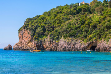 Naklejka premium Seascape – lagoon with turquoise water, mountain with caves and cliffs, green trees, blooming bushes, rocks in a blue water, colorful cruise touristic boats. Corfu Island, Greece. 