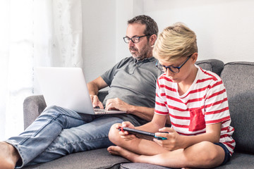 Young boy playing with a video game console while his dad is working with laptop, sitting on a sofa at home. Father and son, generations in comparison. New technology, communication, lifestyle concept