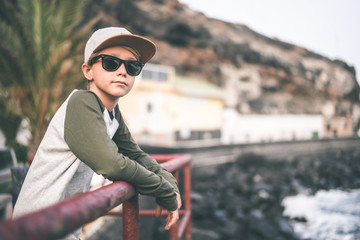 Beautiful smiling teen relaxing leaning against a railing near the sea, looking away with sun glasses and trendy hat. Young boy enjoying last summer days in front of the ocean. Youth, positive concept