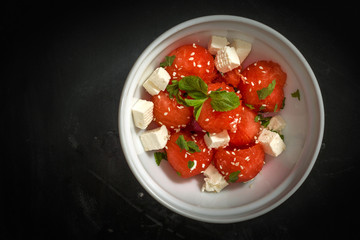 Salad of watermelon balls, feta cheese and mint, sprinkled with sesame seeds in a white plate on a black background.