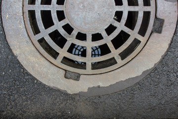 old sewer manhole in gray asphalt and reflection in the water. rough surface texture