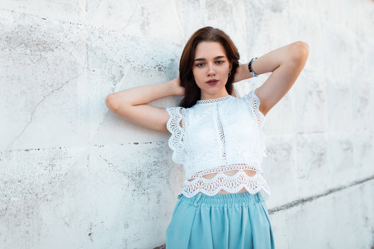 European cute young brunette woman in fashionable white lace blouse in stylish blue pants posing outdoors near a vintage wall. Pretty elegant girl relaxes outdoors on a summer day. New collection.
