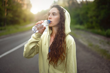 Beautiful young Caucasian girl  standing on asphalt forest road Drinks water from plastic bottle after jogging