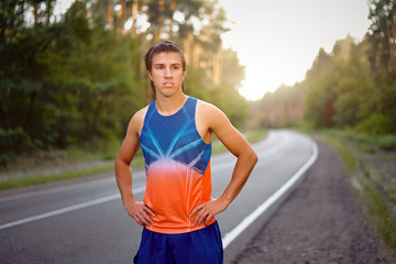 Young curly blonde Caucasian man standing on asphalt forest road looking at camera before jogging.