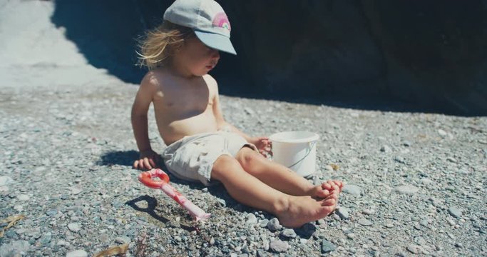 Little toddler sitting on a rocky beach with a bucket and spade