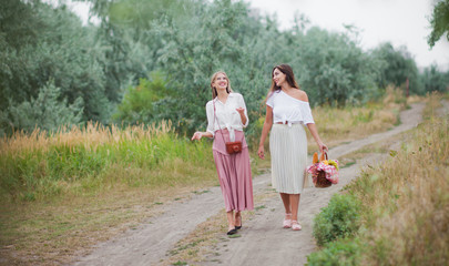 Two cheerful young women in retro style clothes are walking along the landing with a picnic basket and talking. Joint walk in nature
