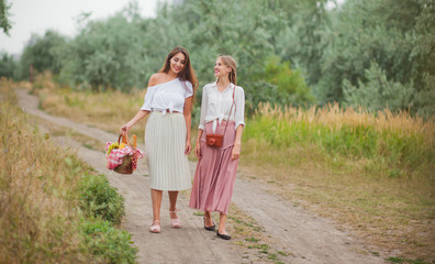 Naklejka na ściany i meble Two cheerful young women in retro style clothes are walking along the landing with a picnic basket and talking. Joint walk in nature