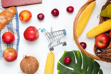 Buying food for a picnic. Mini shopping trolley with fruits and basket on a white background. Monstera leaf.