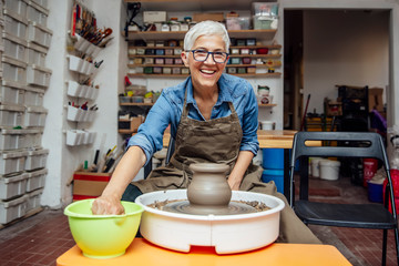 Senior female potter working on pottery wheel while sitting  in her workshop - obrazy, fototapety, plakaty