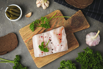 Fototapeta na wymiar Board with pork fatback, garlic, parsley, bread and bowl of pickles on grey stone background, flat lay