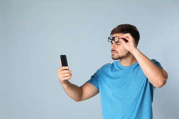 Young man with vision problems using smartphone on grey background, space for text