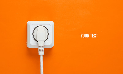 White cable plugged into power outlet on orange wall background with copy space