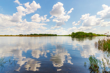 The beautiful sunny landscape with the calm river and fluffy clouds that reflected in the water surface