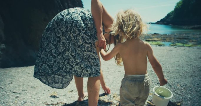 Young mother and toddler walking on beach with bucket, she picks something up