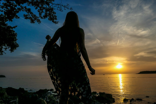 A girl in a maxi dress enjoying the sunset by a stony beach. The sun sets over the horizon. The sun beams reflecting in the calm sea waters. There is an island on the side.Trees growing on the shore.