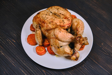 Fried with carrot chicken on black wooden background. Whole grilled chicken with golden crust on a plate. Festive dinner with baked juicy bird.