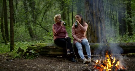 two hikers women are drinking beer on halt in forest in evening near campfire