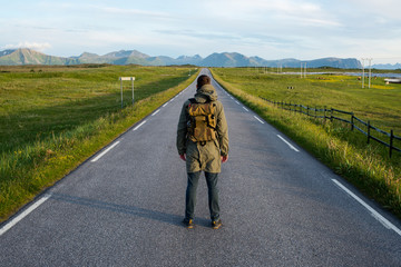 Fototapeta premium Man with backpack walking on an empty road. Amazing landscape, mountain and ocean. Scenic view. New way. Enjoy the moment, relaxation. Wanderlust. Travel, adventure, lifestyle. Explore North Norway