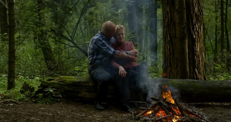 loving aged spouses are embracing sitting near fire in forest, admiring by flaming