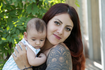 A young happy mother lovingly holds in her arms her 2-month-old son, a newborn, cute baby. Happy motherhood. Girl with a tattoo on her arm.