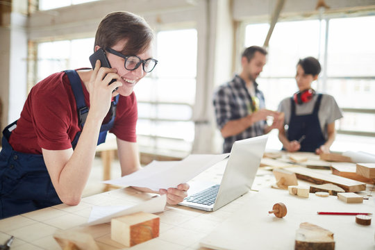 Portrait of modern carpenters working at woodworking factory, focus on smiling young man speaking by phone and using laptop in foreground, copy space