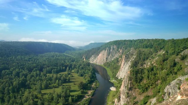 Aerial forward: Flight from a cliff at sunny summer morning of the picturesque nature of the Ural mountains, surrounded by a green forest, river flows at the bottom 