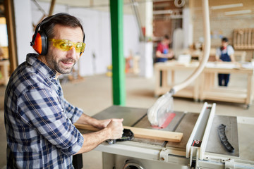 Side view portrait of mature carpenter smiling at camera while polishing wood in joinery workshop, copy space
