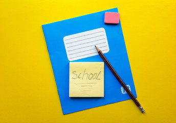 Blue notebook and textbook copy butt for school subjects, eraser, pencil, paper clip on a yellow background. The word school is written in pencil. Flat lay, copy space, top view, place for text.