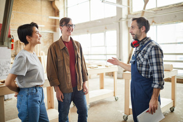 Fototapeta premium Portrait of mature carpenter talking to young people in workshop giving them tour of production, copy space
