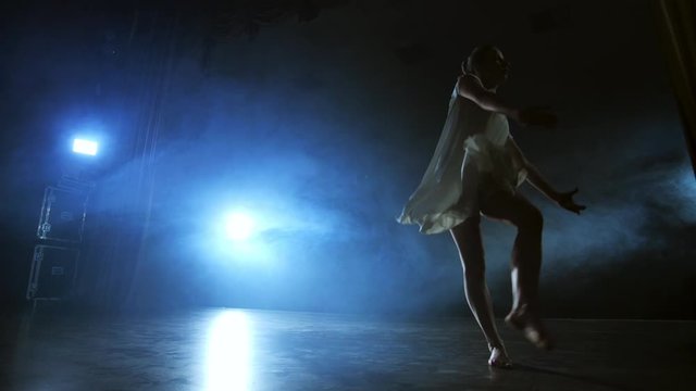 Modern dance woman in a white dress dances a modern ballet, jumps, makes rotation on the stage with smoke in the blue spotlights.