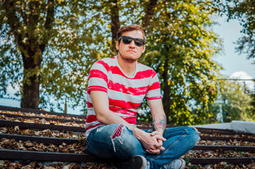 A young handsome man in casual clothes is sitting on the steps in the park and looking around.