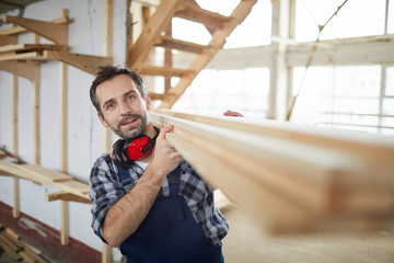 Waist up portrait of mature carpenter looking at camera while carrying wooden boards in industrial workshop, copy space