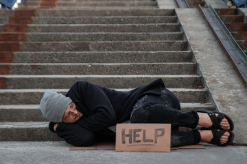 A man, homeless, a man sleeping on a cold floor in the street with a Help sign. Concept of a homeless person, social problem, addict, poverty, despair.
