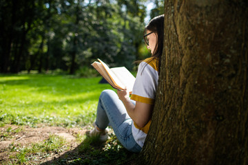 Smiling young woman in jeans reading book resting against park tree view from behind