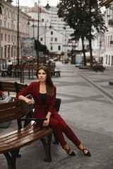 Naklejka premium Fashionable and young pretty woman, in a cherry smart suit, sitting on the bench and relaxing outdoors. Stylish brunette model girl posing on a square and looking aside. Concept of city life and style
