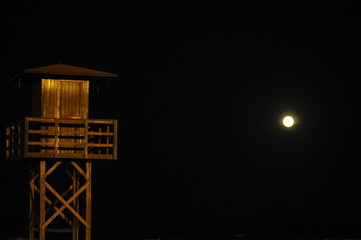 Caseta de guardavidas hecha de madera en la costa de la una playa española en horario nocturno, la luz de la luna deslumbrante en un costado, cielo azul oscuro, azul marino