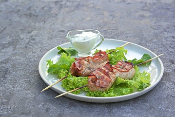 Grilled pork with bacon on wooden skewers, on a white ceramic tray on a gray concrete background. Served with lettuce. Barbecue. Selective focus.
