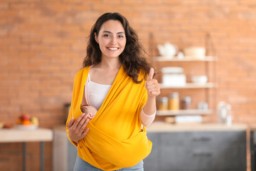 Young mother with little baby in sling showing thumb-up gesture at home