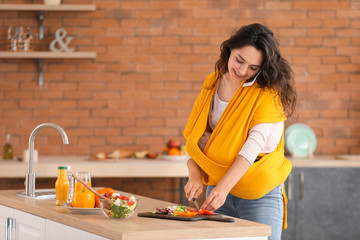 Young mother with little baby in sling and mobile phone preparing vegetable salad at home