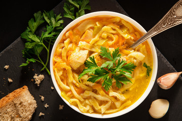 An overhead photo of a plate of chicken and noodles soup, shot from above on a dark rustic texture with a spoon, slices of bread, a celery branch, and garlic