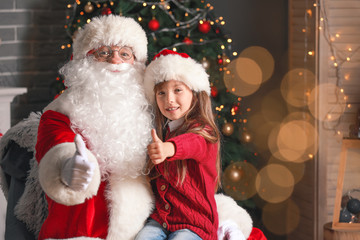 Fototapeta na wymiar Santa Claus and little girl showing thumb-up in room decorated for Christmas